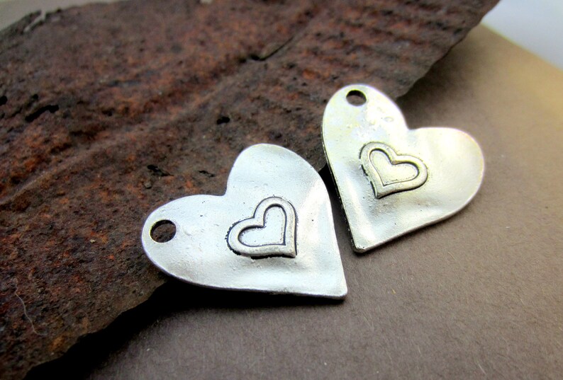 May include: Two silver heart-shaped charms with a smaller heart design in the center. The charms have a hole at the top for attaching to jewelry or other crafts.