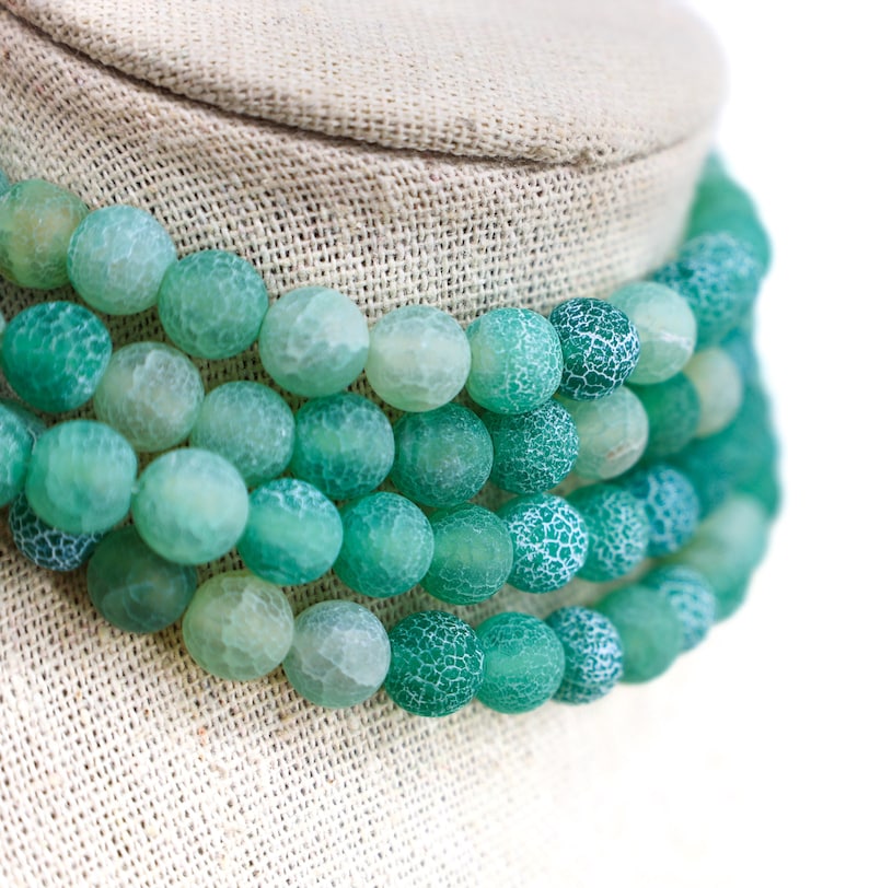 May include: A layered necklace featuring large, round beads in shades of green and teal. The beads have a textured, crackled appearance, creating a unique visual effect. The necklace is displayed on a neutral-toned fabric.