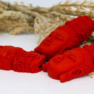 May include: Three red Buddha head sculptures with closed eyes and detailed carved features. The sculptures are arranged on a white surface with dried plants in the background. The heads are a vibrant red color.