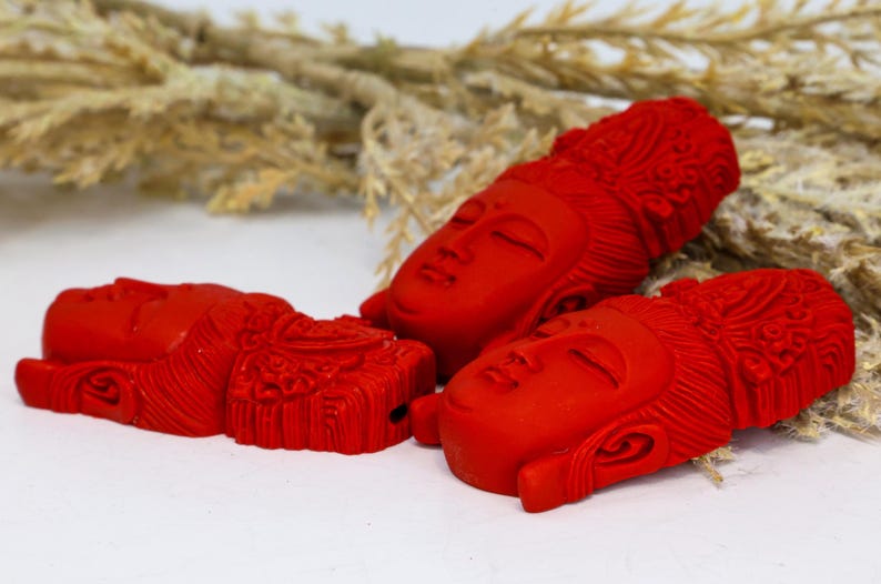 May include: Three red Buddha head sculptures with closed eyes and detailed carved features. The sculptures are arranged on a white surface with dried plants in the background. The heads are a vibrant red color.
