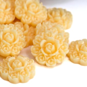 以下が含まれることがあります： 淡い黄色の樹脂製のバラの形をした装飾品のコレクション。詳細な花の模様が一緒に集まっており、一部は重なっています。背景は白で、花の繊細なディテールを際立たせています。