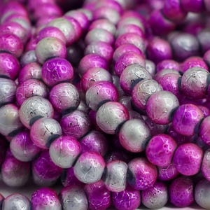 May include: Close-up of a collection of round beads in shades of purple and gray. The beads have a marbled appearance, with the purple hues dominating and the gray adding contrast. The beads are strung together, suggesting they are for jewelry making.