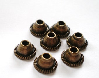 May include: Seven antique bronze-colored metal bead caps. Each cap has a conical shape with a textured, rope-like border. The caps are arranged on a white background, showcasing their detailed design.