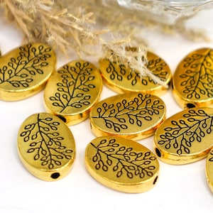 May include: Gold-colored oval beads with a black leaf design. The beads are arranged on a white surface, with dried plants in the background. These beads are suitable for jewelry making and crafting.