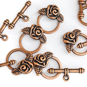 May include: Antique copper-toned metal toggle clasps with a rose design. Each clasp features a round ring with a raised rose and leaves, and a straight bar with a rounded end.