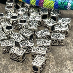 Puede incluir: Una colección de cuentas cuadradas plateadas con un diseño floral y espiral detallado. Cada cuenta tiene un agujero en el centro. Las cuentas están dispersas sobre una superficie de madera, con un textil tejido colorido en el fondo.