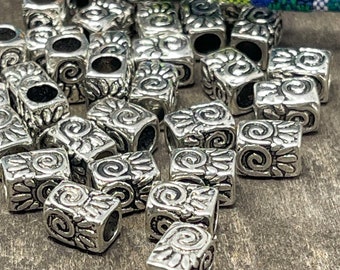 Peut inclure: Collection de perles carrées argentées avec un motif floral et en spirale détaillé. Chaque perle a un trou au centre. Les perles sont dispersées sur une surface en bois, avec un textile tissé coloré en arrière-plan.