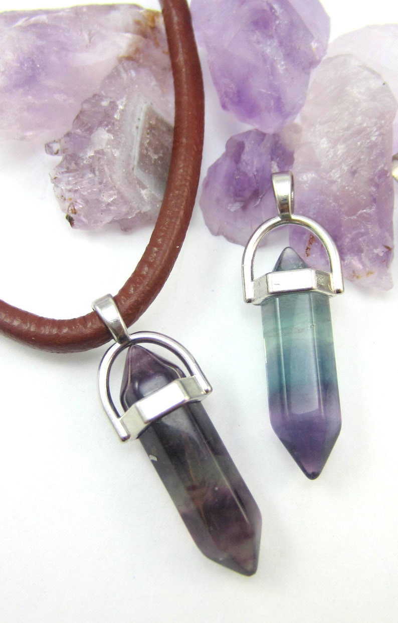 May include: Two silver and crystal point pendants on a brown leather cord. The pendants are shaped like a point and have a silver ring around the top. The crystals are purple and green.