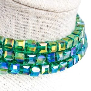 May include: A close-up of a green and iridescent crystal necklace. The necklace is made of square-cut crystals in shades of green, blue, and yellow. The necklace is displayed on a white mannequin head.