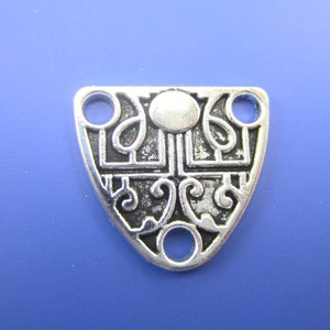 May include: Silver-coloured metal pendant with an ornate, shield-like design. The pendant features a central oval shape, geometric squares, and swirling patterns. It has three holes for stringing or attaching. The background is a solid blue.