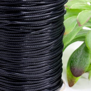 May include: A spool of tightly wound black waxed cord. The cord is a solid black color and appears to be made of a durable material. The spool is sitting on a white surface with green leaves in the background.