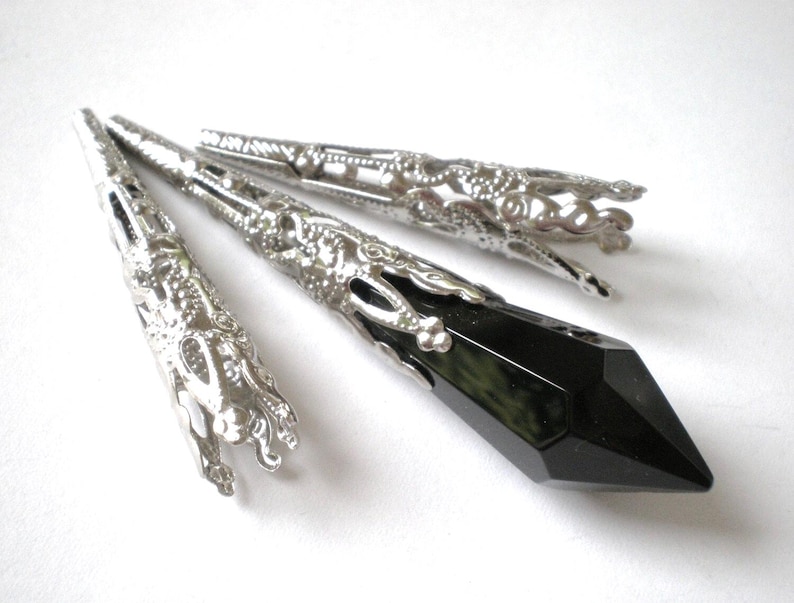 May include: Silver-toned metal jewelry findings with intricate filigree designs. A faceted black crystal pendant is featured, along with three decorative metal caps. The findings are displayed against a white background.