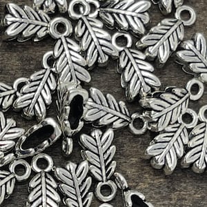 May include: A pile of silver-toned metal leaf-shaped pendant charms with a loop at the top. The charms are scattered on a wooden surface.
