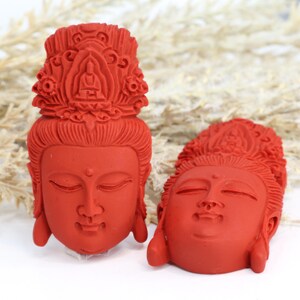 May include: Two red Buddha head sculptures. The sculptures have closed eyes and detailed headdresses. The heads are facing different directions. The background is white with some dried plants.