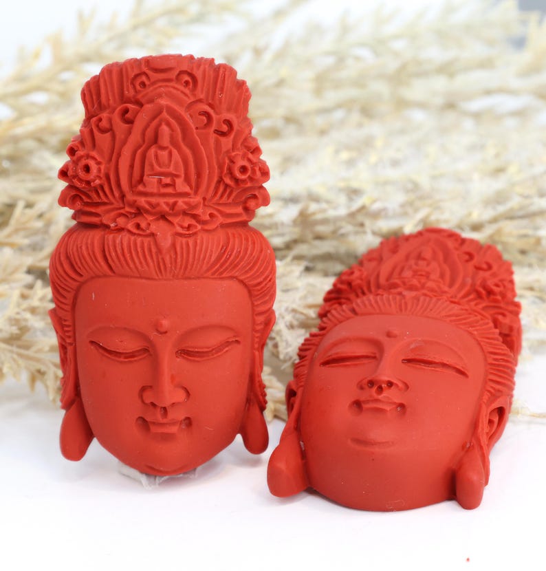 May include: Two red Buddha head sculptures. The sculptures have closed eyes and detailed headdresses. The heads are facing different directions. The background is white with some dried plants.