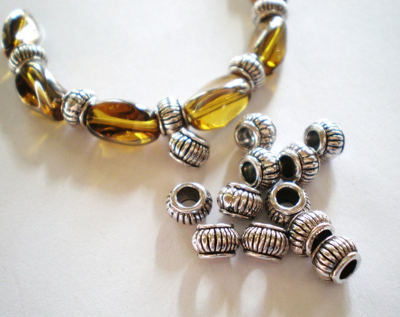 May include: A bracelet with amber-colored glass beads and silver-toned metal accents. Several silver-toned metal beads are scattered nearby. The beads have a ribbed design and are cylindrical in shape. The bracelet is on a white surface.