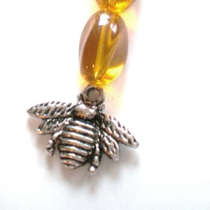 May include: A close-up of a silver-colored bee charm attached to a strand of amber-colored oval beads. The bee charm has detailed wings and body segments. The beads are translucent and have a glossy finish.