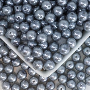 May include: A close-up shot of numerous gray pearl beads, some spilling out of a white, square-shaped container. The beads have a glossy finish and are scattered across the surface. The image showcases the texture and color of the beads.