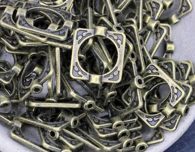 K&ouml;nnte beinhalten: Ein Haufen antiker, bronzefarbener Metallbeschlagteile. Jedes Teil hat einen rechteckigen Rahmen mit einem zentralen Kreis und dekorativen bl&uuml;tenartigen Mustern. Die Beschl&auml;ge haben eine Vintage-&Auml;sthetik, geeignet f&uuml;r Bastel- oder Restaurierungsprojekte.