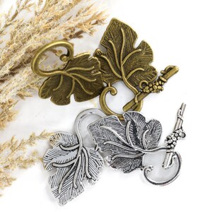 May include: Two toggle clasps shaped like grape leaves and vines, one in an antique brass finish and one in a silver tone. Each clasp consists of a leaf-shaped toggle bar and a circular ring with a small cluster of grapes attached to the side.