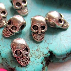 K&ouml;nnte beinhalten: Nahaufnahme von silbernen Totenkopfperlen auf t&uuml;rkisfarbenem Hintergrund. Die Perlen sind klein und detailliert, mit einem realistischen Totenkopfdesign.
