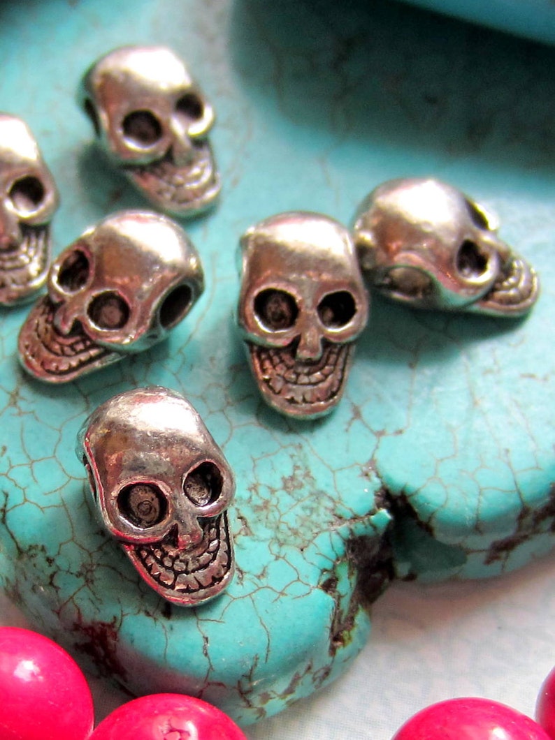 K&ouml;nnte beinhalten: Nahaufnahme von silbernen Totenkopfperlen auf t&uuml;rkisfarbenem Hintergrund. Die Perlen sind klein und detailliert, mit einem realistischen Totenkopfdesign.