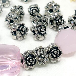 May include: Close-up of jewellery components featuring silver-coloured rose-shaped beads and light pink faceted and rectangular beads. The beads are arranged in a pattern, suggesting a bracelet or necklace design. The background is a soft white.