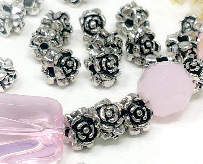May include: Close-up of jewellery components featuring silver-coloured rose-shaped beads and light pink faceted and rectangular beads. The beads are arranged in a pattern, suggesting a bracelet or necklace design. The background is a soft white.