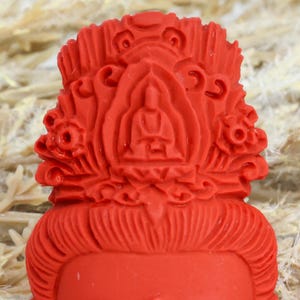 May include: A close-up of a red Buddha statue head. The statue features intricate details, including a serene facial expression, ornate headdress, and a seated Buddha figure at the center. The overall design is symmetrical and detailed.