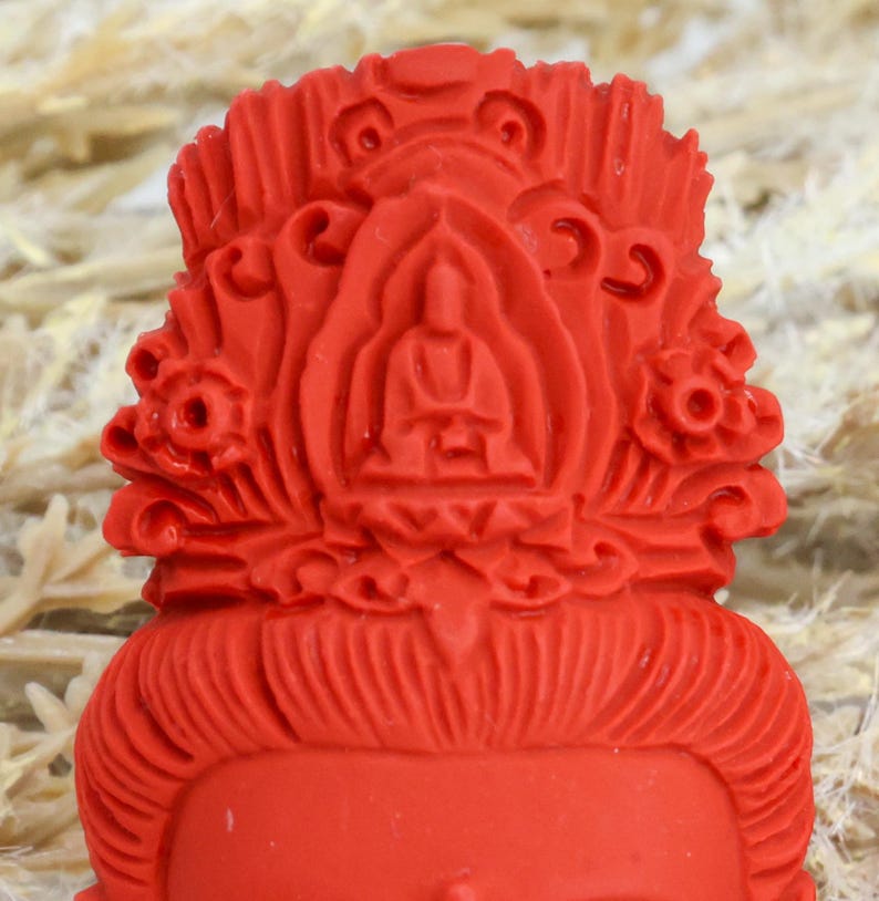 May include: A close-up of a red Buddha statue head. The statue features intricate details, including a serene facial expression, ornate headdress, and a seated Buddha figure at the center. The overall design is symmetrical and detailed.