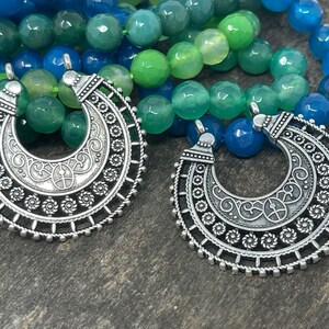 May include: Two silver crescent moon pendants with intricate designs hang from a multi-colored beaded necklace. The necklace is made of green and blue agate beads.