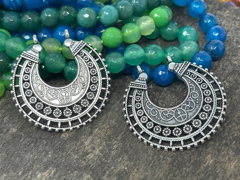 May include: Two silver crescent moon pendants with intricate designs hang from a multi-colored beaded necklace. The necklace is made of green and blue agate beads.