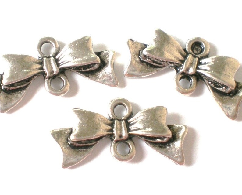 May include: Three silver-toned metal bow charms with two holes each. The bows are detailed with a textured surface and have a three-dimensional appearance.