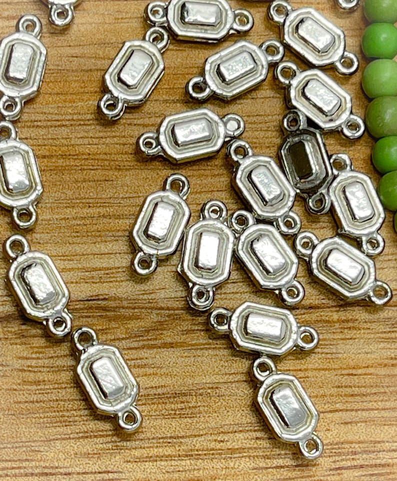 May include: A group of silver-toned metal connector charms with a rectangular shape and a decorative border. The charms have two loops on either side for attaching to jewelry.