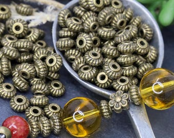 Può includere: Primo piano di un mucchio di perline in metallo dorato antico con un design a festoni. Le perline sono in un cucchiaio di metallo e alcune sono infilate su una collana con perline di vetro ambrato.