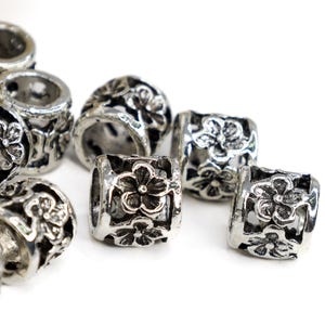May include: Silver metal beads with a floral design. The beads are cylindrical and have a hole in the center. They are perfect for making jewelry or other crafts.