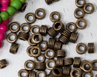 Può includere: Una collezione di piccole perline metalliche cilindriche con una finitura in bronzo antico e un motivo a griglia testurizzato sull'esterno. Un filo parziale di perline di legno rotonde rosa brillante, verde lime e giallo è visibile nell'angolo in alto a sinistra.