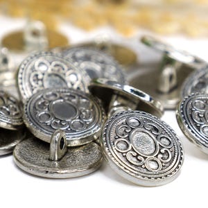 May include: A collection of antique silver-colored buttons. Each button has a decorative, circular design with an ornate pattern. The buttons are piled together, showing their detailed craftsmanship.