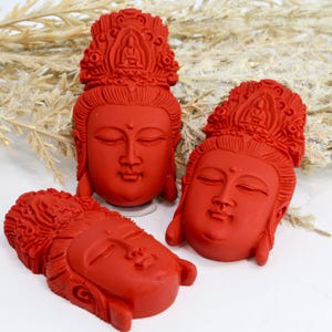 May include: Three red Buddha head sculptures with closed eyes and ornate headdresses. The sculptures are arranged on a white surface with dried plants in the background. The heads are carved with detailed facial features.