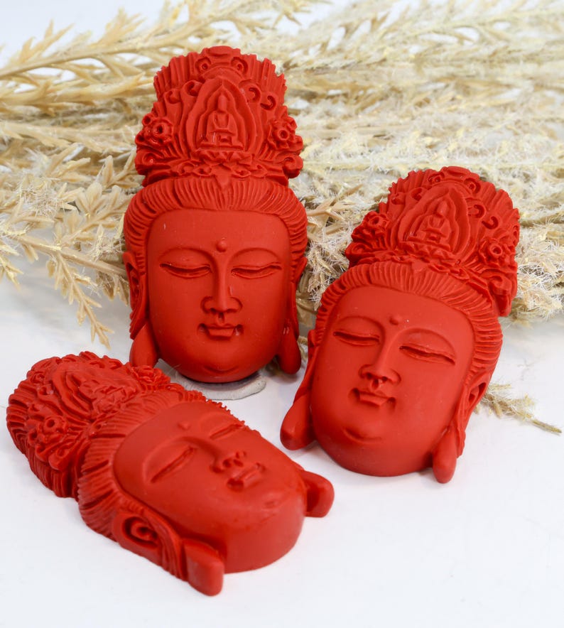 May include: Three red Buddha head sculptures with closed eyes and ornate headdresses. The sculptures are arranged on a white surface with dried plants in the background. The heads are carved with detailed facial features.