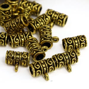 May include: A collection of antique gold-colored metal beads. The beads are cylindrical with intricate scrollwork and small loops for stringing. The beads are scattered on a white surface, showcasing their detailed design and vintage aesthetic.