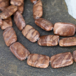 May include: A strand of brown and tan rectangular gemstone beads. The beads have a marbled pattern and are strung together with a clear thread.
