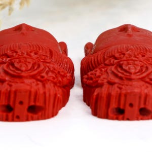 May include: Two red, carved figurines with detailed facial features and ornate headdresses. The figurines are symmetrical, with a smooth, glossy finish. The background is white.