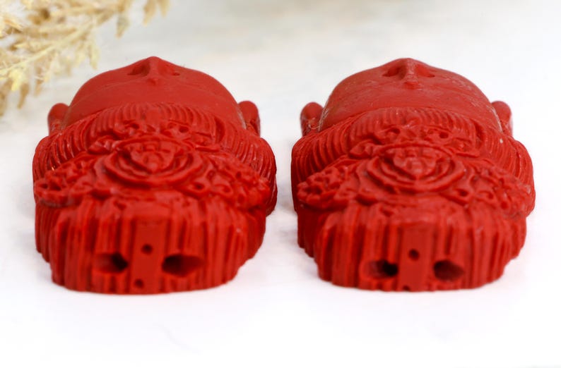 May include: Two red, carved figurines with detailed facial features and ornate headdresses. The figurines are symmetrical, with a smooth, glossy finish. The background is white.