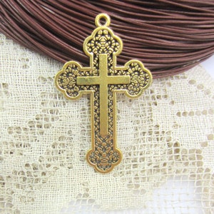 May include: A gold-colored cross pendant with ornate floral detailing. The cross features a classic Christian design with a vertical and horizontal bar. The pendant has a loop at the top for attaching to a chain or cord. The background includes brown leather cord and lace.