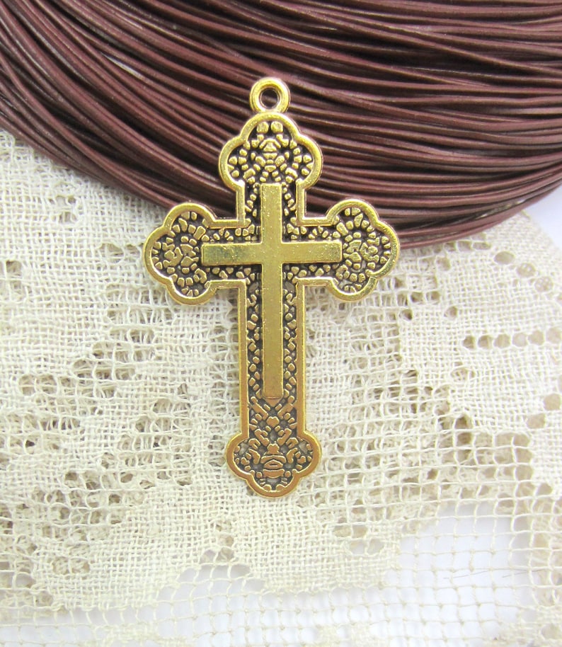 May include: A gold-colored cross pendant with ornate floral detailing. The cross features a classic Christian design with a vertical and horizontal bar. The pendant has a loop at the top for attaching to a chain or cord. The background includes brown leather cord and lace.