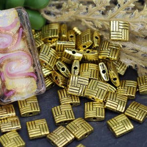 以下が含まれることがあります： 織り模様の入った金色の正方形の金属ビーズの山。ビーズの中央には糸を通すための穴があります。