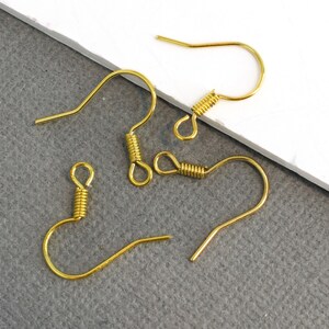 May include: Close-up of several gold-colored earring hooks. The hooks have a curved design with a looped end and a coiled spring detail. The hooks are made of a shiny, metallic material, and are displayed against a neutral background.