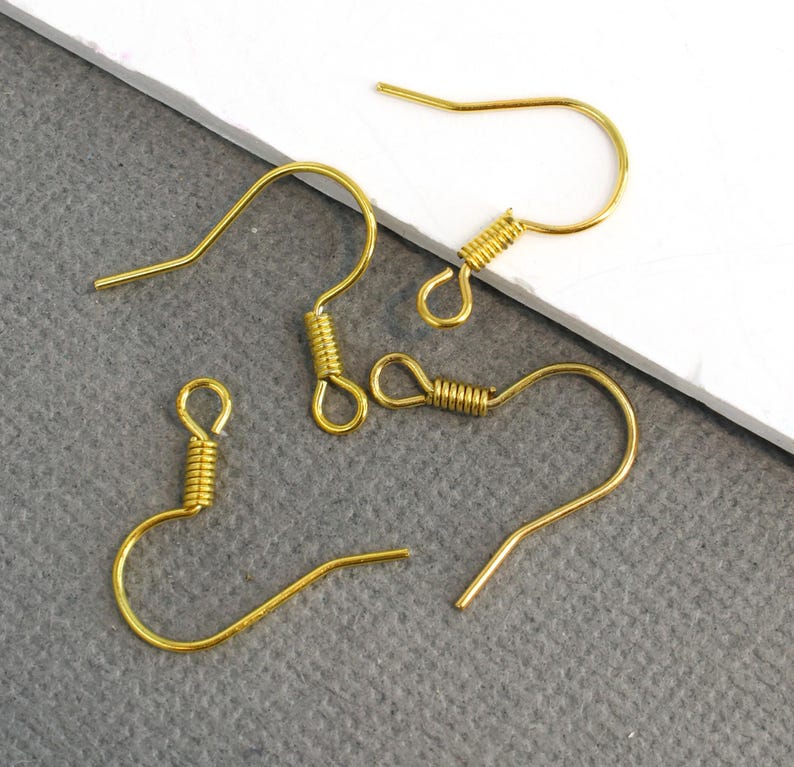 May include: Close-up of several gold-colored earring hooks. The hooks have a curved design with a looped end and a coiled spring detail. The hooks are made of a shiny, metallic material, and are displayed against a neutral background.