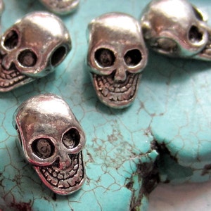 K&ouml;nnte beinhalten: Eine Gruppe silberner Totenkopfperlen mit detaillierten Gesichtsz&uuml;gen. Die Perlen sind auf einer t&uuml;rkisfarbenen Oberfl&auml;che angeordnet.